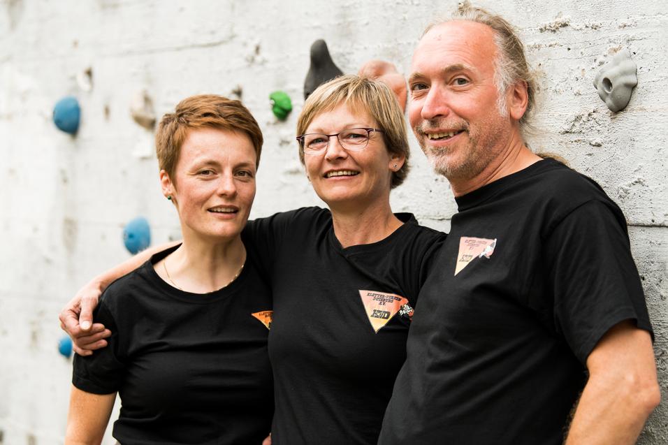 Drei Personen in schwarzen T-Shirts lächeln vor einer Kletterwand mit bunten Griffen.
